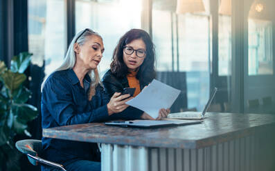 Zwei Geschäftsfrauen, die an einem Tisch sitzen und zusammen arbeiten. Eine leitende Angestellte zeigt ihr Telefon und ihre Papiere einem jungen Kollegen im Büro. - JLPSF22707