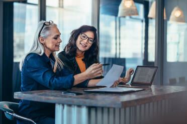 Two women analyzing documents while sitting on a table in office. Woman executives at work in office discussing some paperwork. - JLPSF22710