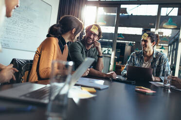 Business team sitting around a table in office lobby with sticky notes on their forehead having a brainstorming meeting. Corporate business people sitting in board room and discussing. - JLPSF22751