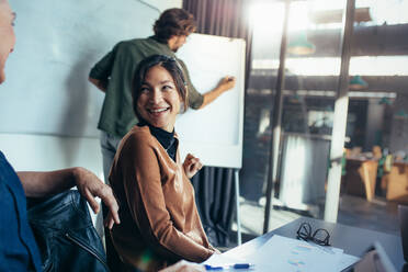 Smiling asian woman talking with coworker sitting by in business presentation. Business presentation in modern office board room. - JLPSF22754