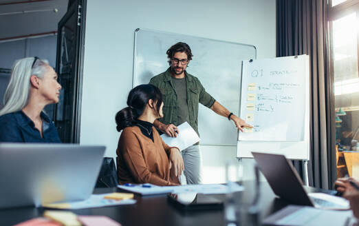 Young man standing by flipchart and giving presentation over new business strategy for the first financial quarter. Young man giving business presentation to colleagues sitting around table in conference room. - JLPSF22755