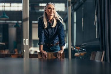 Portrait of successful businesswoman standing leaning to a chair in meeting room and looking away. Senior female entrepreneur in conference room. - JLPSF22769