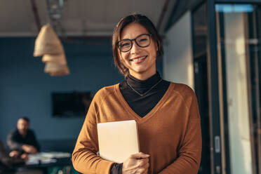 Portrait of happy business woman holding a laptop standing in office with colleagues working in background. Smiling woman in casuals in office. - JLPSF22777