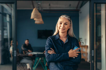 Portrait of smiling mature woman standing outside meeting room with team discussing work in background. Smiling senior business woman at office. - JLPSF22779