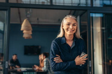 Portrait of smiling senior woman standing in office doorway with her arms crossed. Beautiful female executive at office with people meeting in background. - JLPSF22780