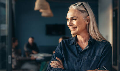 Close up of senior business woman standing in office with her arms crossed and looking away smiling. Mature female in office with team meeting in background. - JLPSF22781