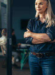 Vertical shot senior business woman standing outside meeting room with colleagues discussing in background. Mature business woman in casuals at office. - JLPSF22786
