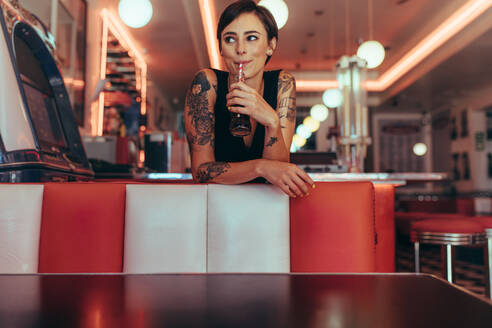 Woman with tattooed hands drinking soft drink with a straw at a restaurant. Happy woman standing in a cafe drinking a soft drink looking away. - JLPSF22791