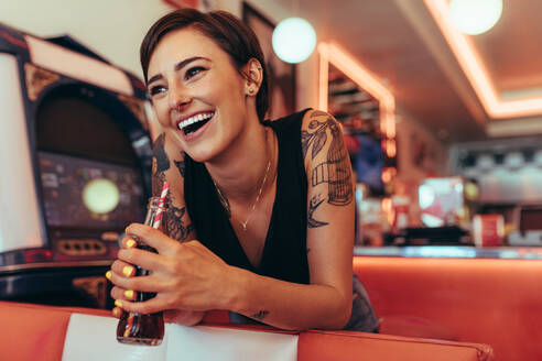 Lächelnde Frau mit tätowierten Armen, die ein Erfrischungsgetränk trinkt und neben einer Spielhalle steht. Glückliche Frau, die in einem Diner steht und ein Erfrischungsgetränk hält. - JLPSF22793