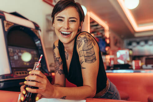 Smiling woman with tattooed arms standing in a diner. Woman standing beside a gaming machine holding a soft drink. - JLPSF22795