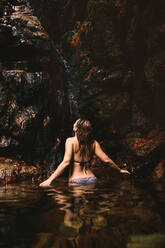 Rear view of woman in bikini taking bath under the waterfall in forest. Female enjoying in a waterfall pond. - JLPSF22798