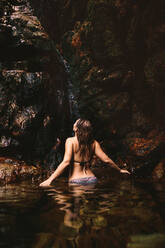 Rückansicht einer Frau im Bikini, die ein Bad unter einem Wasserfall im Wald nimmt. Frau genießt in einem Wasserfallteich. - JLPSF22798
