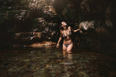 Beautiful young woman in a waterfall pond in forest. |Female wearing bikini standing in the pond by rocks and looking up. - JLPSF22799