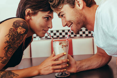Nahaufnahme eines glücklichen Paares, das mit Strohhalmen an einem Milchshake aus einem Glas nippt und sich dabei ansieht. Mann und Frau teilen sich einen Milchshake mit zwei Strohhalmen. - JLPSF22800