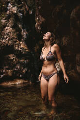 Woman standing inside water pool in the forest and looking up at the view. Female in bikini enjoying the nature. - JLPSF22802