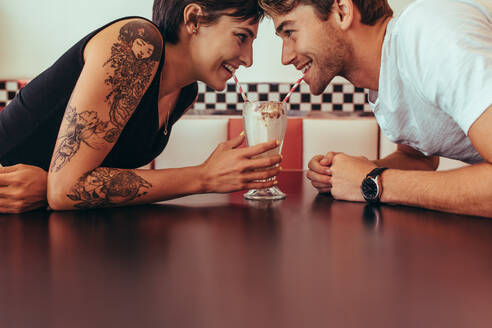 Nahaufnahme eines glücklichen Paares, das sich einen Milchshake mit Strohhalmen teilt und sich in einem Restaurant gegenübersitzt. Romantisches Paar, das an einem Milchshake aus einem Glas nippt und sich dabei ansieht. - JLPSF22803