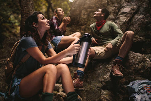 Eine Frau sitzt auf einem Felsen und hält eine Thermoskanne in der Hand, während ihre Freunde hinten im Wald sitzen. Eine Gruppe von Freunden macht eine Pause beim Wandern in der Natur. - JLPSF22804