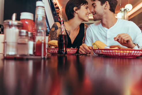 Romantic couple at a diner sharing a french fry holding it in their teeth together and looking at each other. Happy couple dining at a restaurant with food on the table. - JLPSF22809