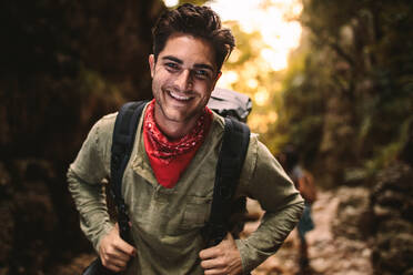 Porträt eines glücklichen jungen Mannes mit Rucksack, der in den Bergen wandert, mit Freunden im Rücken. Der Mann schaut in die Kamera und lächelt beim Wandern in der Natur. - JLPSF22811