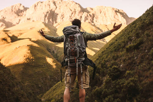 Rückansicht eines jungen Mannes mit Rucksack, der mit ausgestreckten Armen auf einem Felsen steht und auf die Berglandschaft blickt. Männlicher Wanderer steht auf einem Felsen und blickt mit ausgestreckten Armen auf die Berge. - JLPSF22819