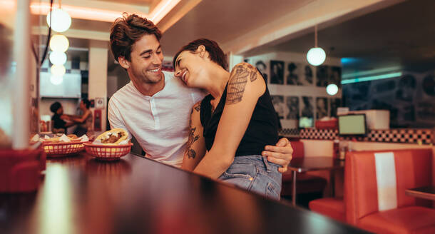 Romantic couple sitting in a restaurant with food on the table. Smiling woman sitting with her boy friend in a restaurant and resting her head on his shoulders. - JLPSF22821