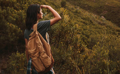 Junge Frau mit Rucksack, die auf einem Bergpfad steht und mit der Hand auf der Stirn in die Ferne blickt. - JLPSF22825