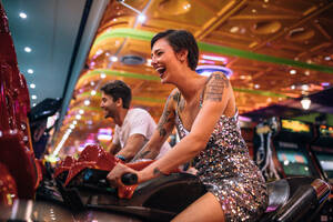 Excited woman playing a racing game sitting on an arcade racing bike. Couple enjoying arcade racing games at a gaming parlour. - JLPSF22840