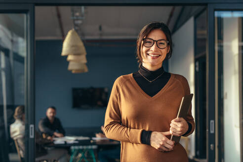 Porträt einer positiven weiblichen Führungskraft, die einen Laptop hält und im Büro steht, mit Kollegen, die im Hintergrund arbeiten. Lächelnde Geschäftsfrau in Freizeitkleidung im Büro. - JLPSF22846