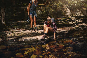 Zwei Wanderinnen an einem Teich in den Bergen: Eine Frau sitzt auf einem Felsen und hält ihre Hand ins Wasser, ihre Freundin steht daneben und schaut zu. - JLPSF22857