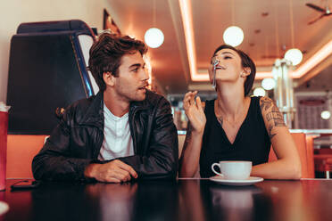 Eine Frau, die sich einen Löffel an die Nase hält, sitzt mit ihrem Freund in einem Diner. Ein Paar sitzt in einem Restaurant mit einem Kaffee auf dem Tisch und hat Spaß. - JLPSF22862