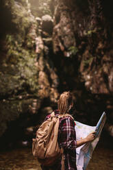 Rückansicht einer jungen Frau mit Rucksack, die eine Wanderkarte in der Hand hält und auf eine Aussicht blickt; Wanderin mit Karte schaut weg. - JLPSF22867