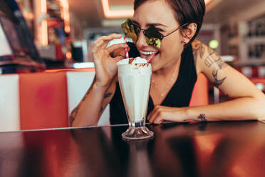 Eine Frau mit einer schicken Brille sitzt in einem Restaurant und genießt ihren Eis-Milchshake. Eine lächelnde Frau leckt den Belag auf dem Milchshake und schaut durch ihre sternförmige Schutzbrille. - JLPSF22869