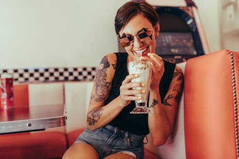 Woman with tattooed arms drinking milkshake sitting at a restaurant. Happy woman in fancy eyewear sitting at a diner drinking milkshake with a straw. - JLPSF22870