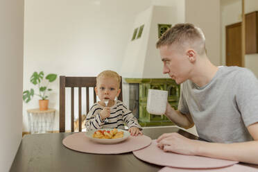 Niedlicher Junge isst Essen mit Vater hält Teetasse am Esstisch - VIVF00111