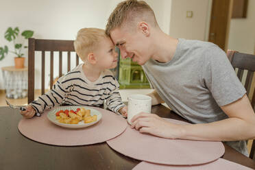 Glücklicher Vater und Sohn, die sich am Esstisch die Stirn berühren - VIVF00112