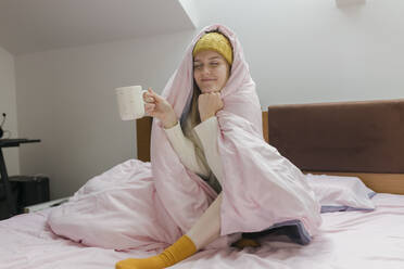 Junge Frau hält Kaffeetasse in Decke eingewickelt auf Bett zu Hause - VIVF00123
