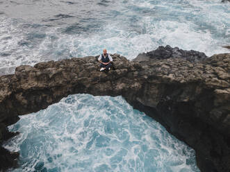 Mature man practicing meditation on rock arch above sea - HMEF01418
