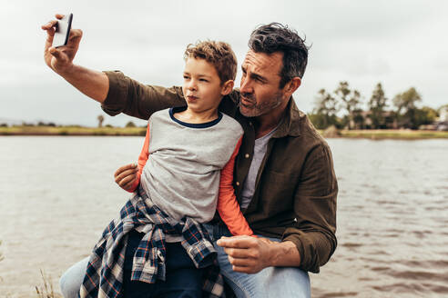 Nahaufnahme von Vater und Sohn, die ein Selfie in der Nähe eines Sees machen. Ein glückliches Kind sitzt auf dem Schoß seines Vaters und schaut auf das Handy, während sein Vater ein Selfie macht. - JLPSF22902