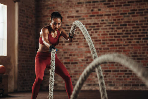 Junge muskulöse Frau trainiert mit Battle Ropes im Fitnessstudio. - JLPSF22910