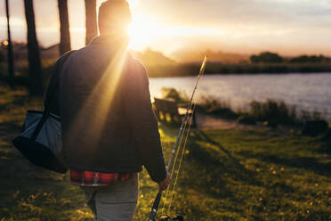 Rückansicht eines Mannes, der mit einer Angelrute auf einen See zugeht. Nahaufnahme eines Mannes, der zum Angeln geht, mit der aufgehenden Sonne im Hintergrund. - JLPSF22913