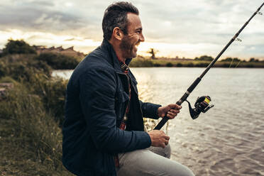 Smiling man sitting beside a lake and fishing. Side view of a happy man sitting near a lake holding a fishing rod. - JLPSF22918