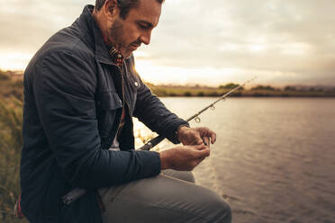 Side view of a man fishing near a lake in his leisure time. Man sitting near a lake fixing a bait to his fishing rod. - JLPSF22921