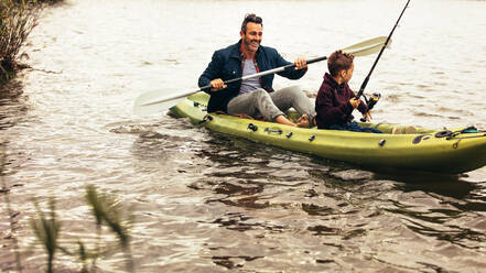 Vater und Sohn gehen in einem Kajak auf einem See angeln. Der Junge sucht mit einer Angelrute nach Fischen im See, während sein Vater das Boot rudert. - JLPSF22925