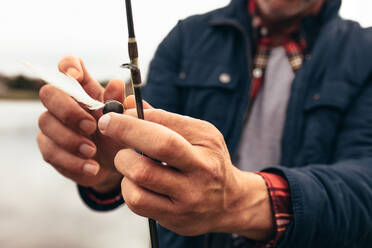 Cropped shot of a man tying a bait to his fishing rod. Close up of hands of a person fixing a bait to a fishing rod to catch fish. - JLPSF22928