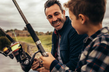 Close up of father and son fishing together sitting near a lake. Smiling man and kid looking at each other enjoying fishing holding fishing rods. - JLPSF22929