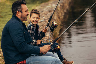 Side view of a man sitting on the banks of a lake and fishing with his kid. Close up of a man winding the reel of his fishing rod while his son looks at him. - JLPSF22931