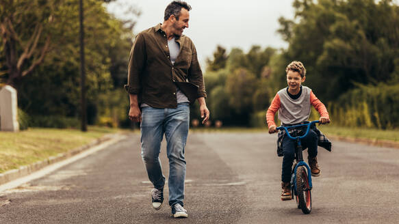 Ein Junge fährt Fahrrad, während sein Vater neben ihm hergeht. Ein Kind lernt mit seinem Vater das Fahrradfahren. - JLPSF22939