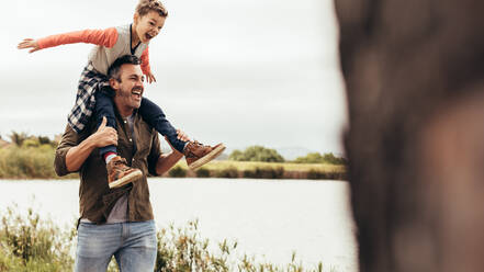 Man walking near a lake with his son sitting on his shoulders. Father and son having fun together enjoying near a lake. - JLPSF22941