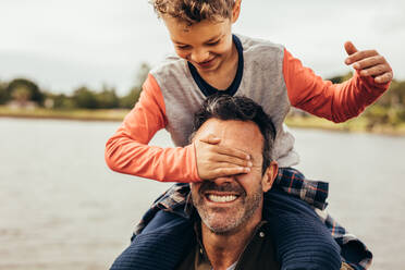Father and son having fun spending time together playing outdoors. Boy closing the eyes of his father sitting on his shoulders. - JLPSF22942