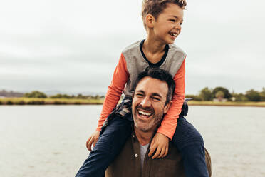 Kid sitting on the shoulders of his father and having fun. Portrait of a smiling man with his kid sitting on his shoulders near a lake. - JLPSF22943